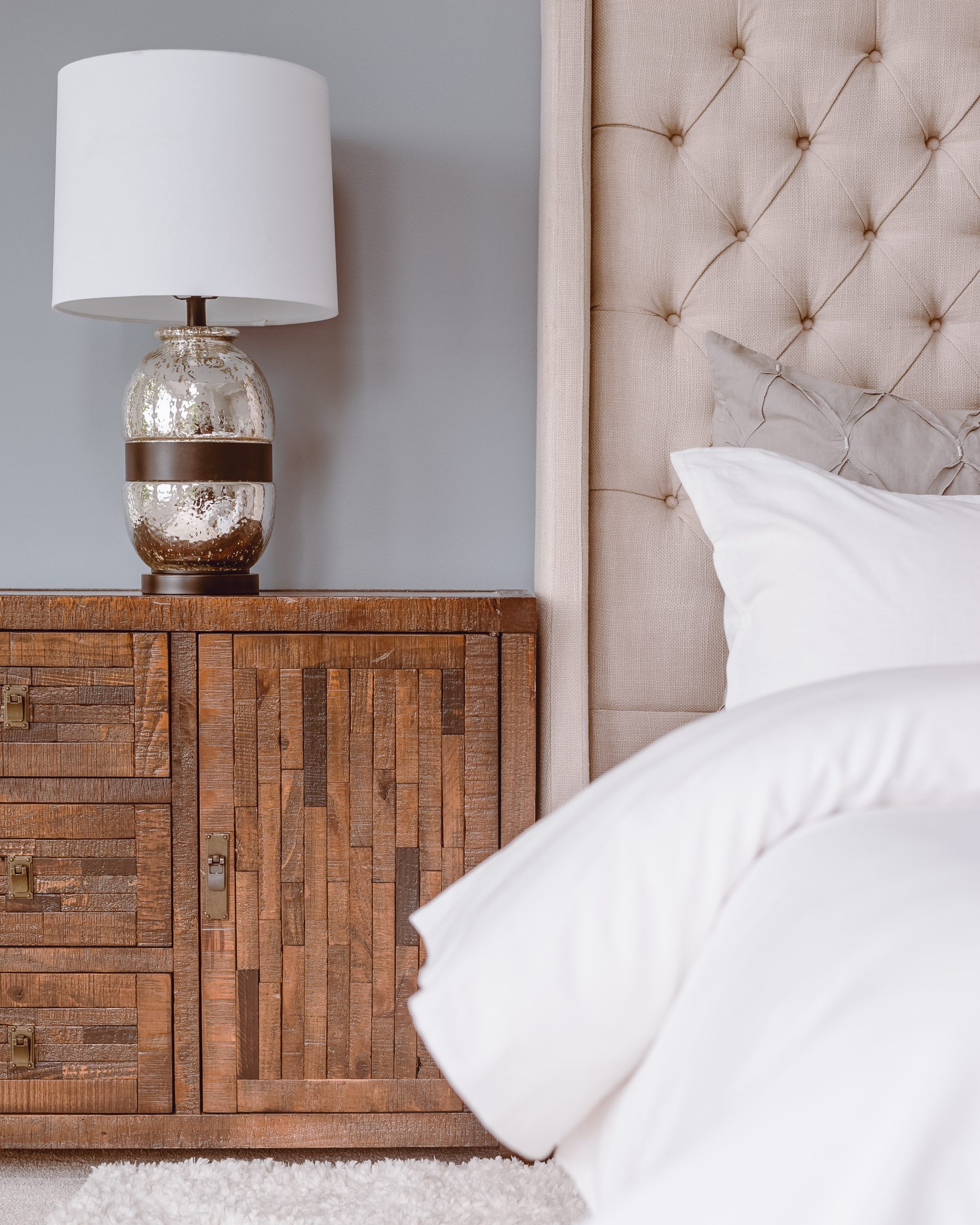 A bed with white pillows and sheets with an adjacent wood-paneled nightstand and table lamp.